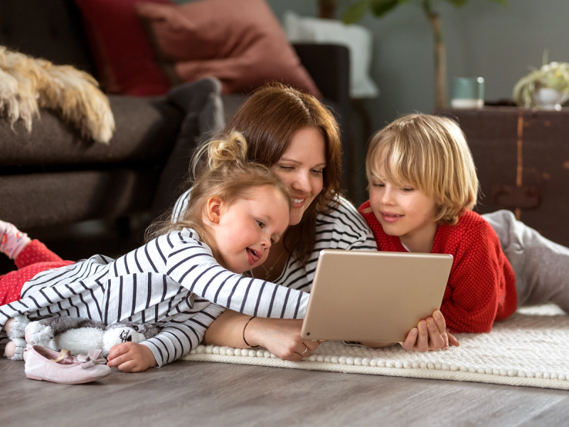 Tre barn ligger på magen på gulvet og ser på en nettbrett sammen i stua, med en voksen i bakgrunnen.