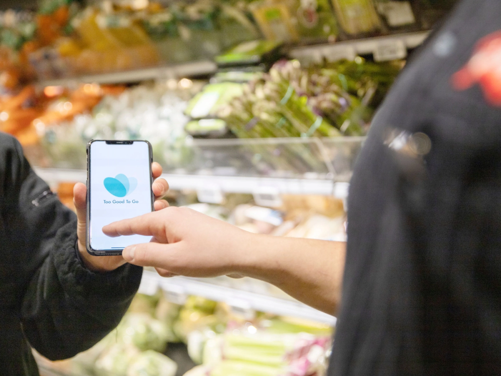 En person viser en mobiltelefon med Too Good To Go-appen foran en grønnsaksavdeling i en butikk.