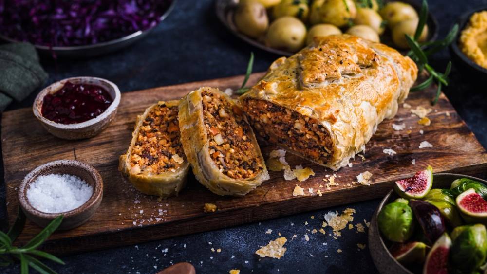 Nøttestek skåret opp på et skjærebrett med tilbehør som syltetøy og salt, omgitt av frukt og urter.