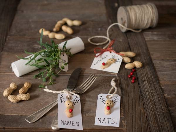 Julekort og navnelapper med reinsdyrmotiv, peanøtter, en liten plante, og bestikk på et treverk, klar til julefeiring.