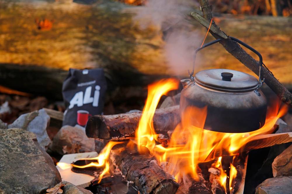 Kjele med bålkaffe som koker over åpen ild med steiner rundt, og en termos i bakgrunnen.