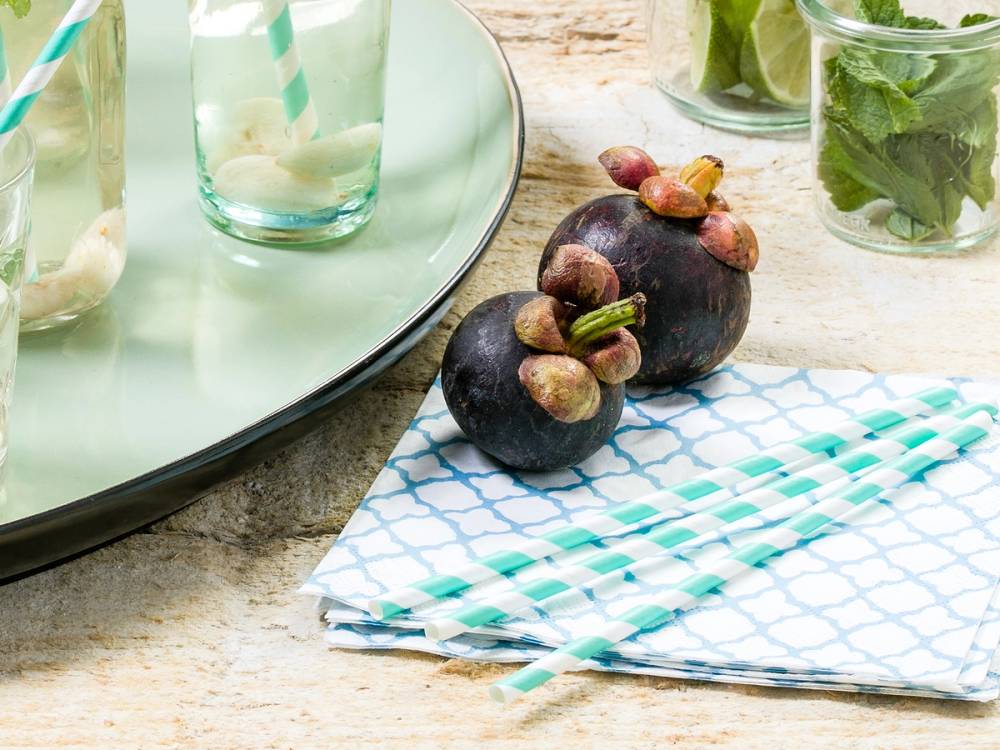 To mangostanfrukter med lilla skall og grønne stilker ligger på et bord ved siden av glass med vann og urter, samt en skål med urter i bakgrunnen.