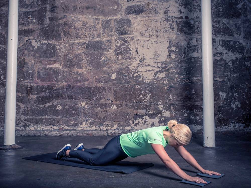 En kvinne i grønn treningstøy gjør en uttøyningsøvelse på en yogamatte mot en murvegg.