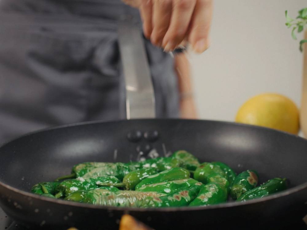 Ferske padron peppers stekes i en panne, med en hånd som holder en pinsett over dem.