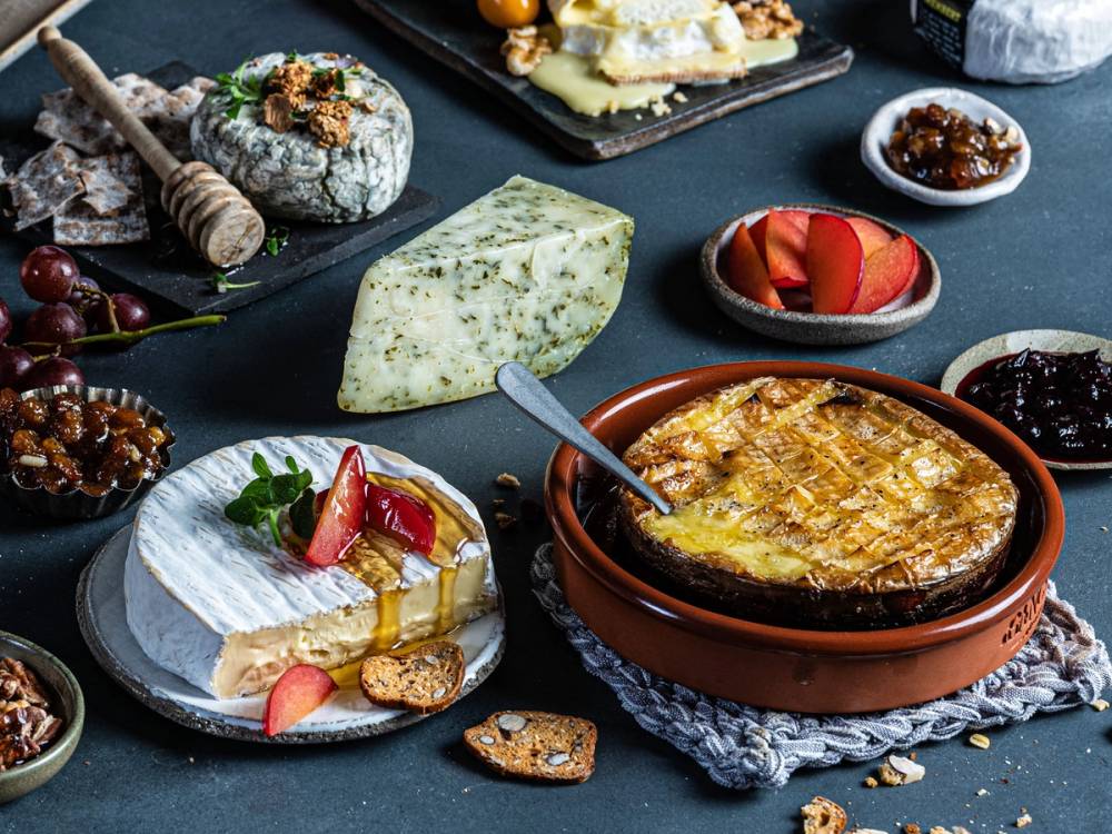 Et ostefat med ulike typer ost, inkludert en blåmuggost, og små skåler med tilbehør som syltetøy og frukt, på et svart bord.