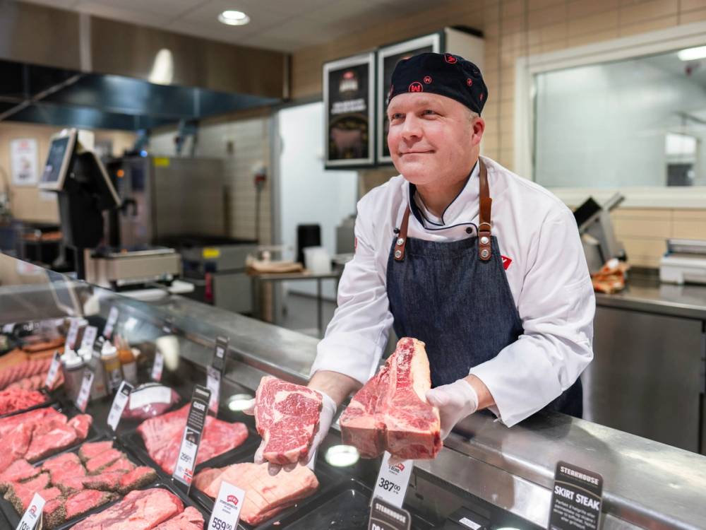 En mann i hvit skjorte og blå forkle står bak en kjøledisk med ulike kjøttstykker i en matbutikk, og holder et stykke kjøtt i hendene.