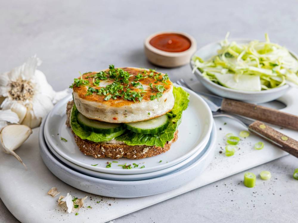 Fiskeburger med stekt fisk, salat og dressing på en tallerken, med en skål med ketchup og en skål med kål i bakgrunnen.