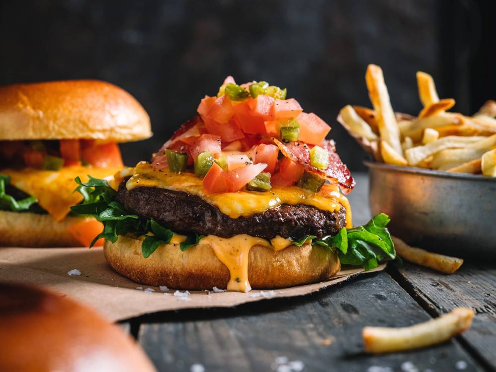 En spicy burger med bacon, tomat, løk, og chipotlemajones, servert med pommes frites på siden.