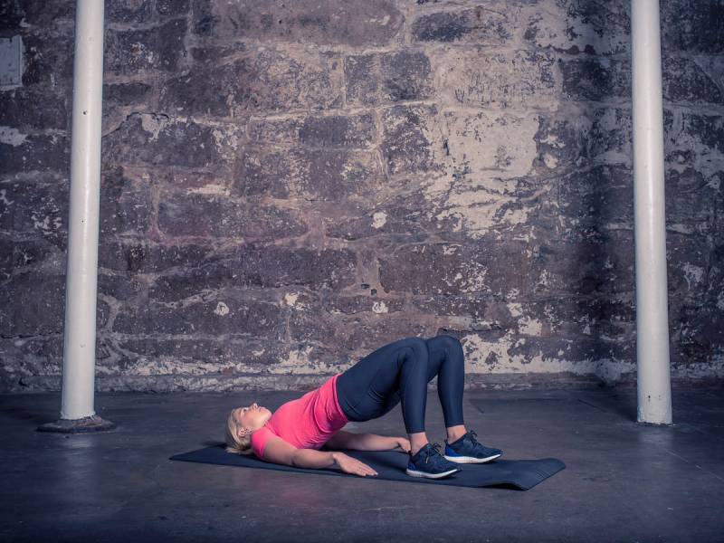 En kvinne utfører en setehev-øvelse på en treningsmatte i et treningsrom med murvegg og stenger på sidene.