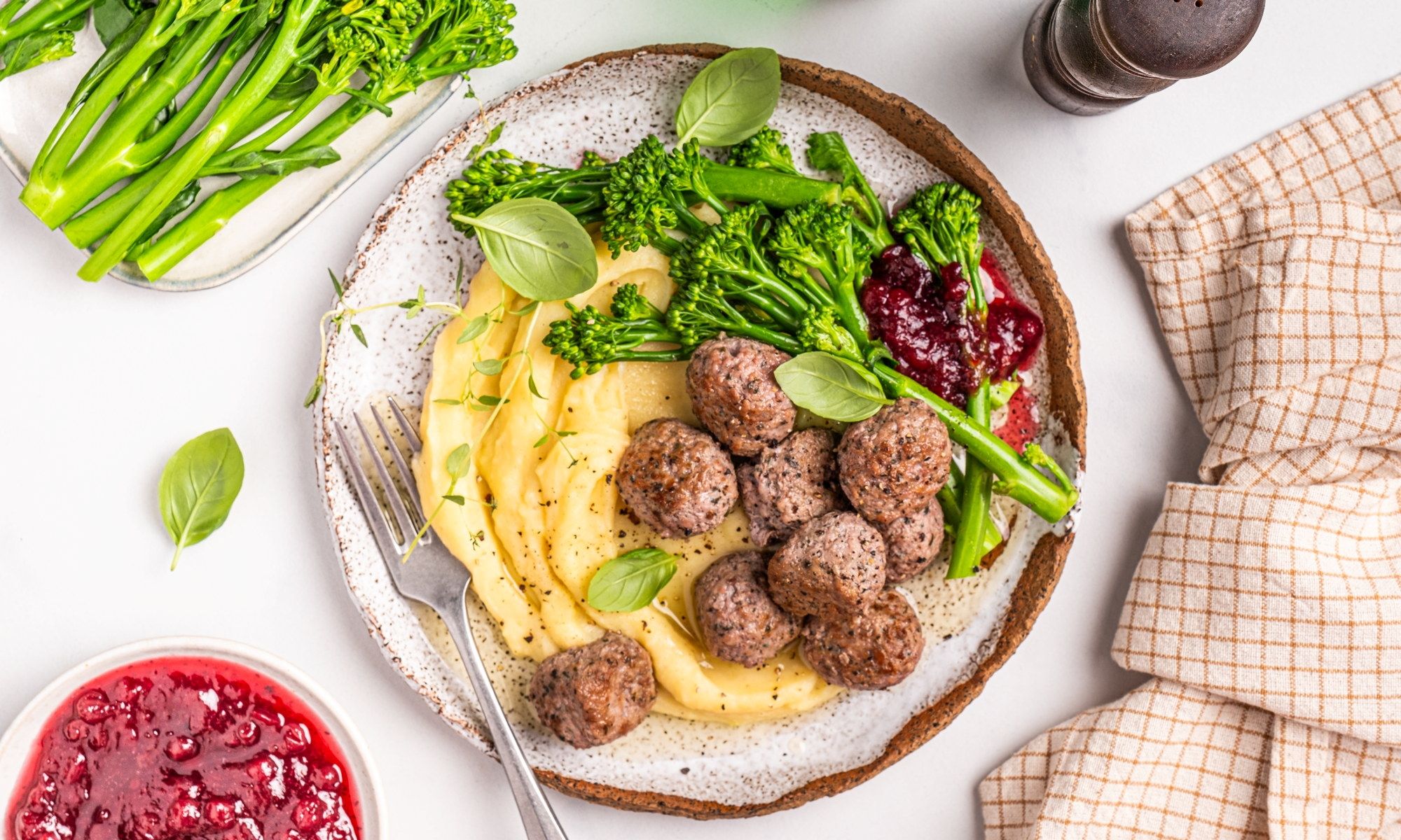 Kjøttboller med potetmos og grønnsaker på en tallerken, omgitt av tilbehør som grønne urter og saus.