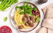 Kjøttboller med potetmos og grønnsaker på en tallerken, omgitt av tilbehør som grønne urter og saus.