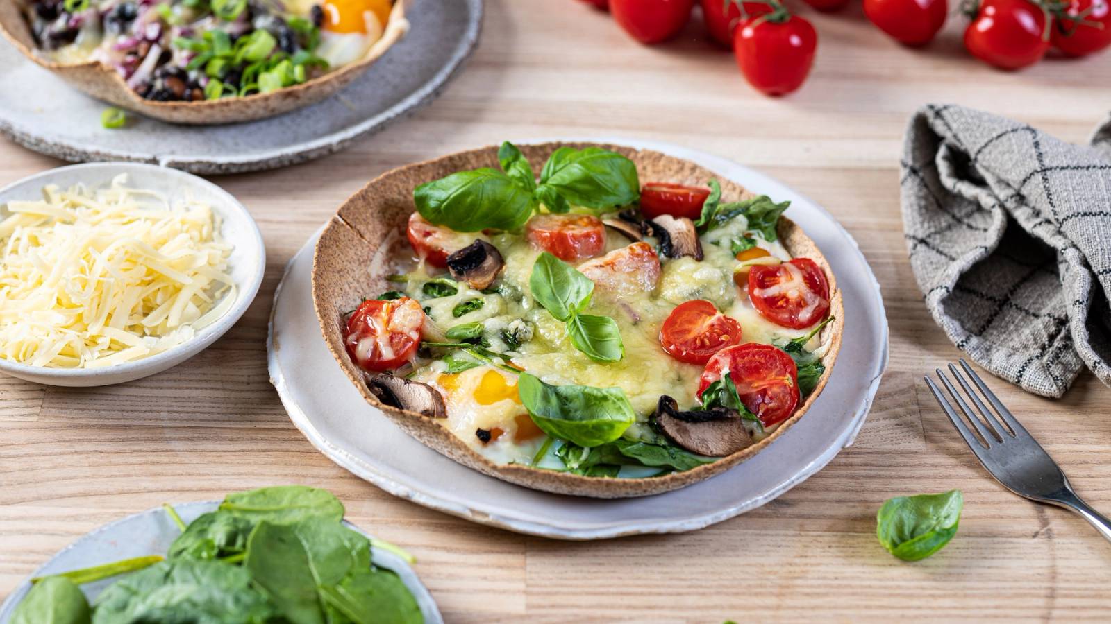 En pizza med tomater, salat og ost på en treplate, omgitt av cherrytomater og en skål med revet ost.