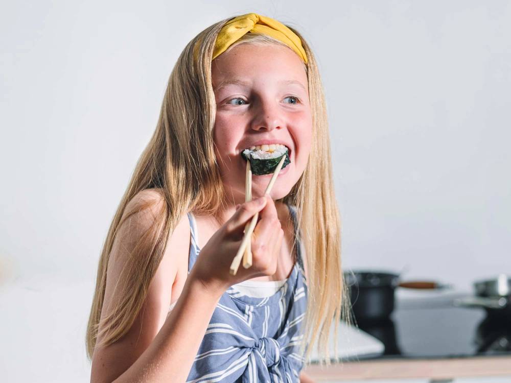 En jente som spiser mat i et kjøkken, med fokus på matglede, og hun har langt blondt hår og en blå og hvit kjole.