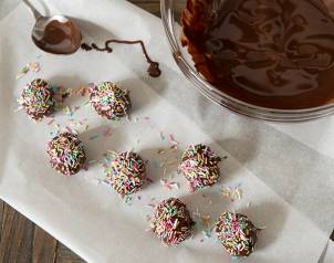 Hjemmelaget påskegodteri med sjokoladekuler dekket av fargerike strøssel, plassert på et hvitt papirark ved siden av en bolle med smeltet sjokolade.