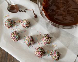 Hjemmelaget påskegodteri med sjokoladekuler dekket av fargerike strøssel, plassert på et hvitt papirark ved siden av en bolle med smeltet sjokolade.