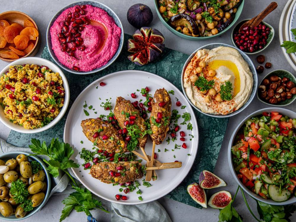 Små retter og dipper med ulike farger og teksturer på et bord, inkludert hummus, salat og frukt, med en tallerken med kjøttspyd i midten.