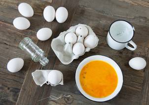 Egg som er blitt knust, med noen hele egg, en liten flaske, en kopp med melk og en skål med eggeplommer på et trebord.