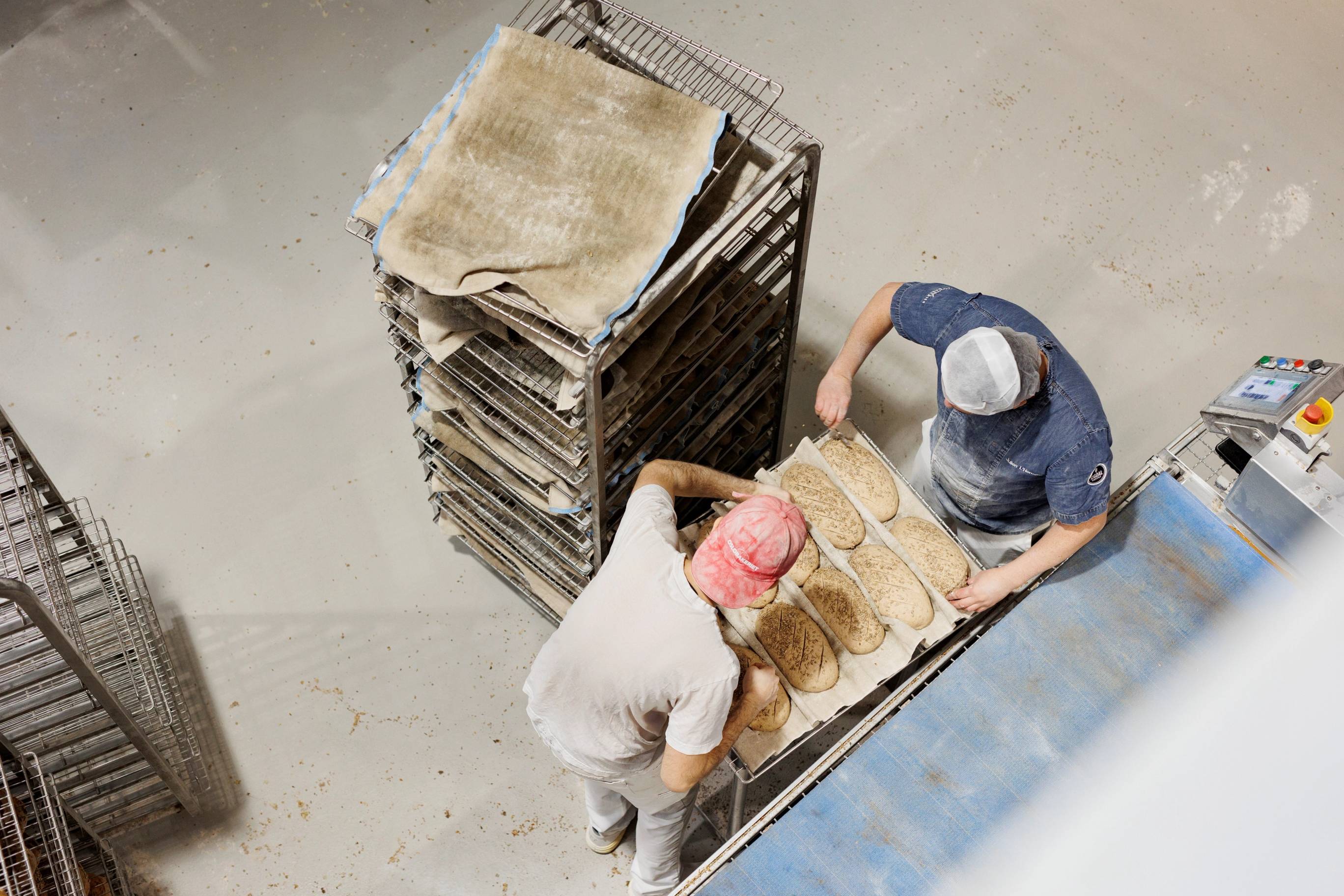 To personer håndterer brød på et bakeri, med en tralle og et stativ med bakevarer, sett ovenfra.