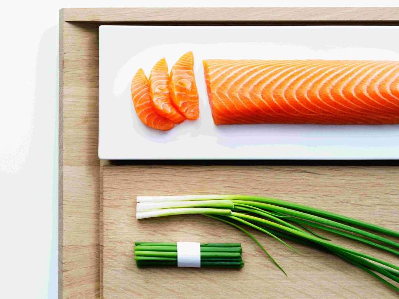 Fersk laksefilet med skiver av sitron og vårløk på et hvitt skjærebrett.