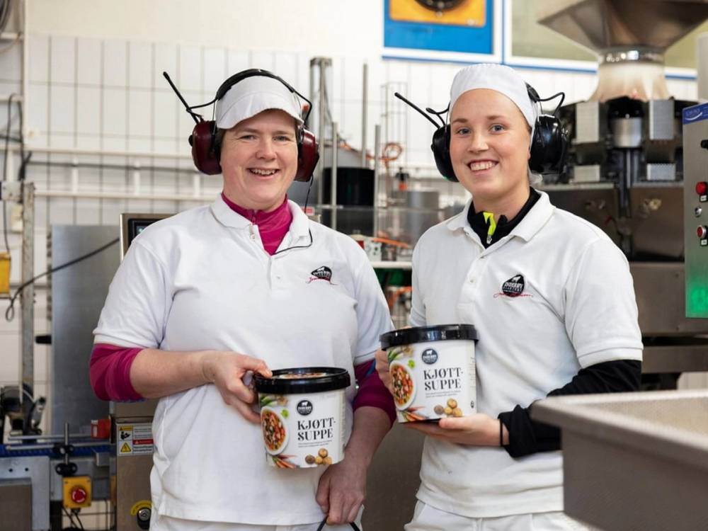 To ansatte i hvite arbeidsklær som står i et kjøkkenmiljø og holder hver sin boks med kjøttsuppe.