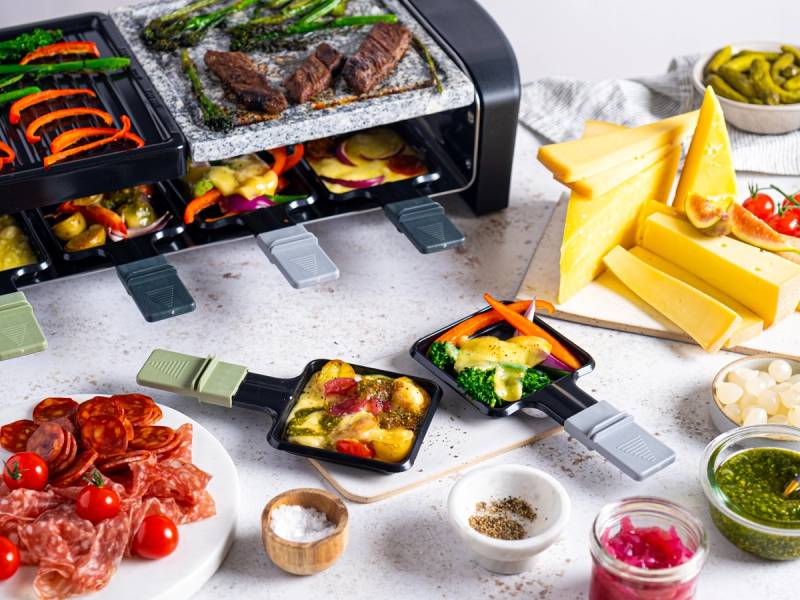 Raclette-plate med smeltet ost, grønnsaker og kjøtt, omgitt av tilbehør som poteter, salat og krydder på et kjøkkenbord.