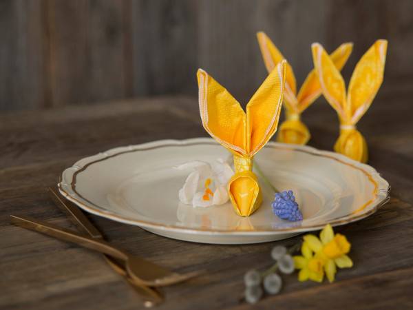 Hareservietter formet som påskeharer på et serveringsfat med blomster og dekorasjoner rundt.