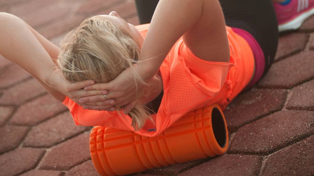 En person ligger på bakken med en massasjerull under ryggen, mens en annen person gir en massasje.