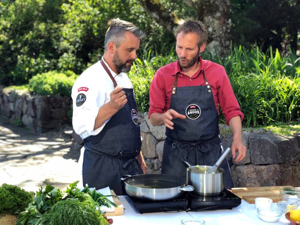 To personer står ute i en hage eller park, og lager mat på en gassgrill, med grønne planter og trær i bakgrunnen.