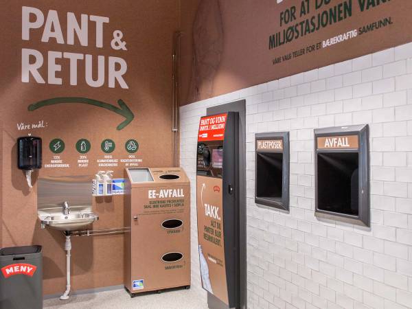 Returpunkt med to avfallscontainere og informasjonsskilt om kildesortering, plassert på en vegg i et offentlig rom.