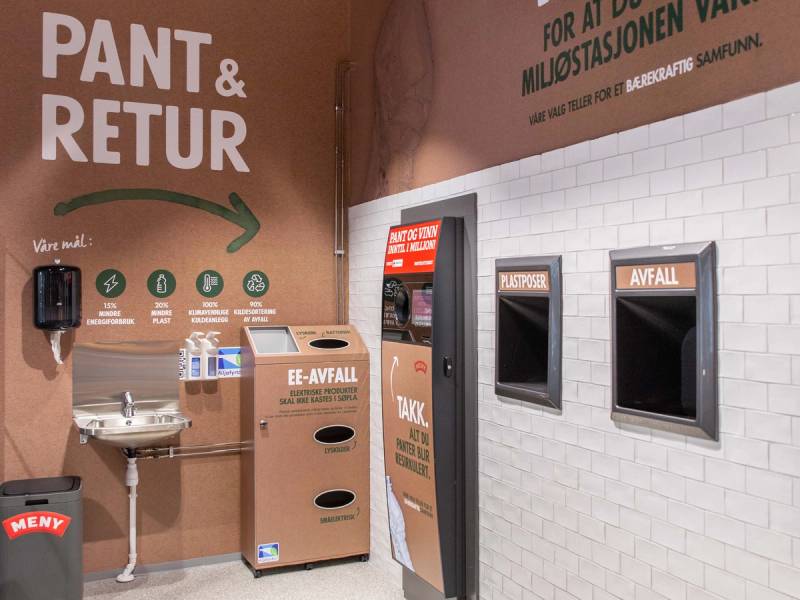 Returpunkt med to avfallscontainere og informasjonsskilt om kildesortering, plassert på en vegg i et offentlig rom.