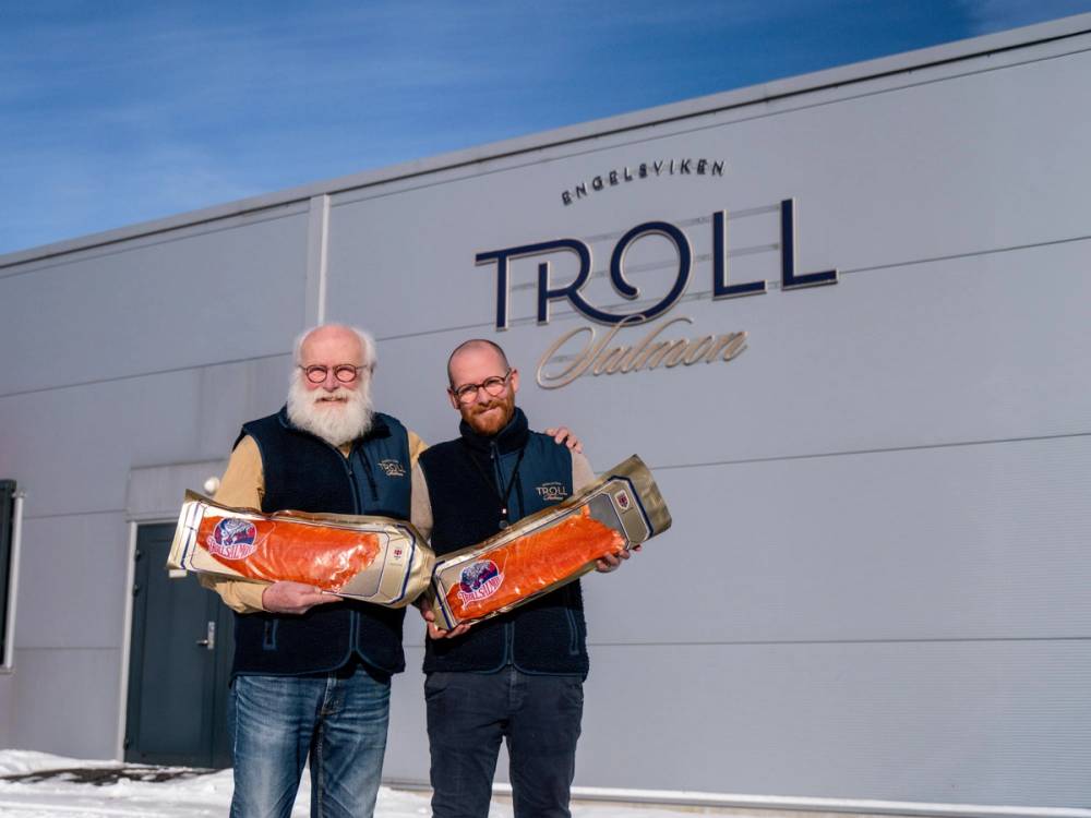 To personer med hver sin pakke røkelaks foran bygg med Troll Salmon-logo.
