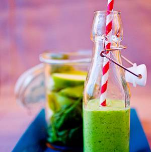 En grønn smoothie i en glassflaske med et rødt og hvitt sugerør, plassert foran en glasskrukke med grønne blader og fruktstykker.