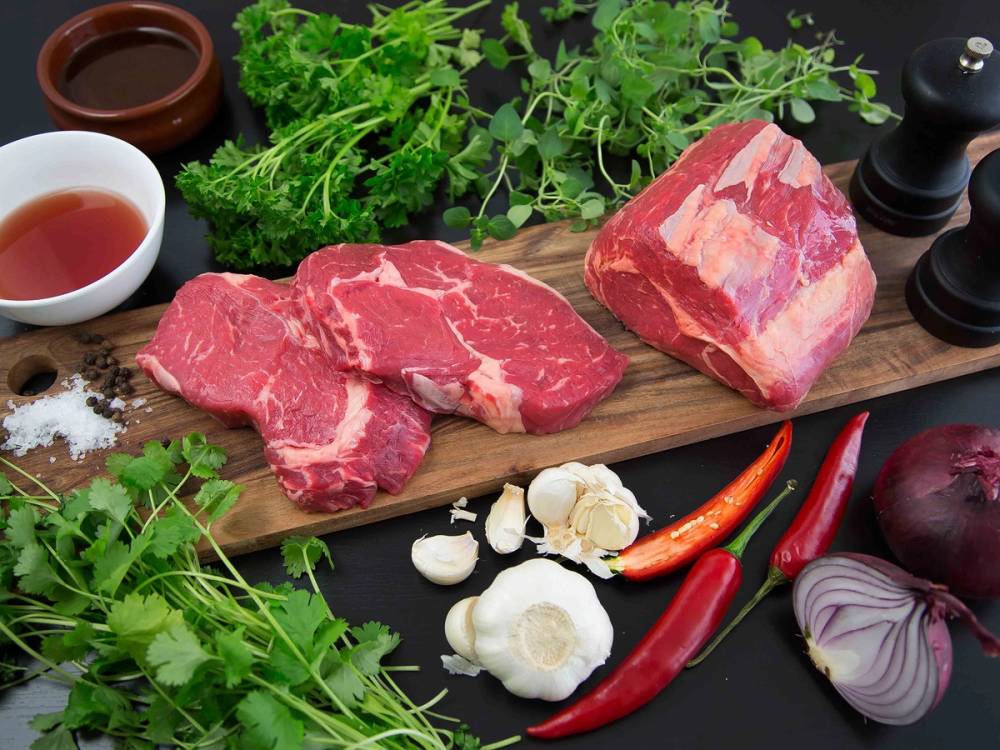 Ulike rå entrecote-biter på et skjærebrett med urter, grønnsaker og krydder rundt.