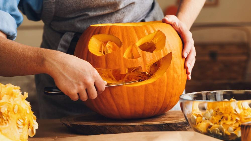 En person skuler ut innmaten av en gresskar for å lage en halloween-dekorasjon, med en bolle med gresskarbiter ved siden av.
