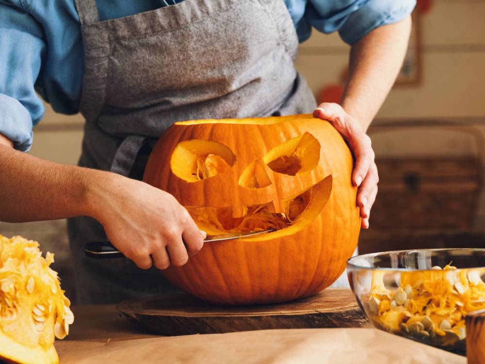 En person skuler ut innmaten av en gresskar for å lage en halloween-dekorasjon, med en bolle med gresskarbiter ved siden av.