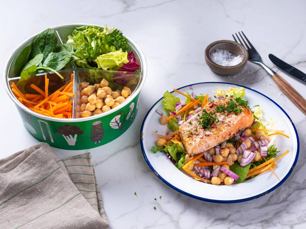 En salat med laks, kikerter, gulrøtter, og blandet grønt på en tallerken, med en liten skål salt og bestikk ved siden av.