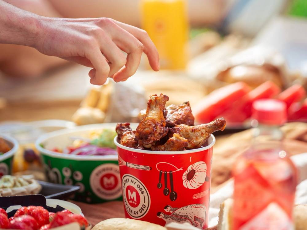 En person plukker opp en kyllingvinge fra en rød kopp med mat, omgitt av ulike matretter og drikker på et bord.