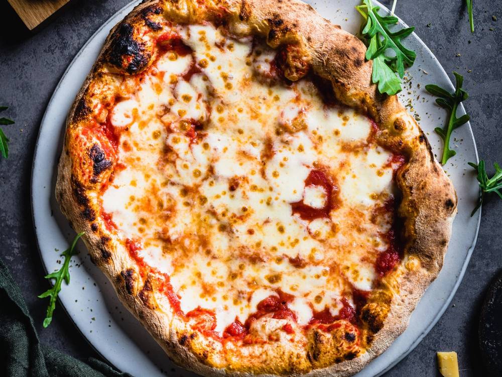 En pizza med ost og tomatsaus på en rund tallerken, med noen grønne urter ved siden av.