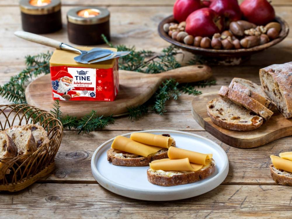 Julebrunost-ny-pakning-2.jpg viser en pakke med brunost på et trefat, med skiver av brunost på brød, omgitt av nøtter, epler og juledekorasjoner på et bord.