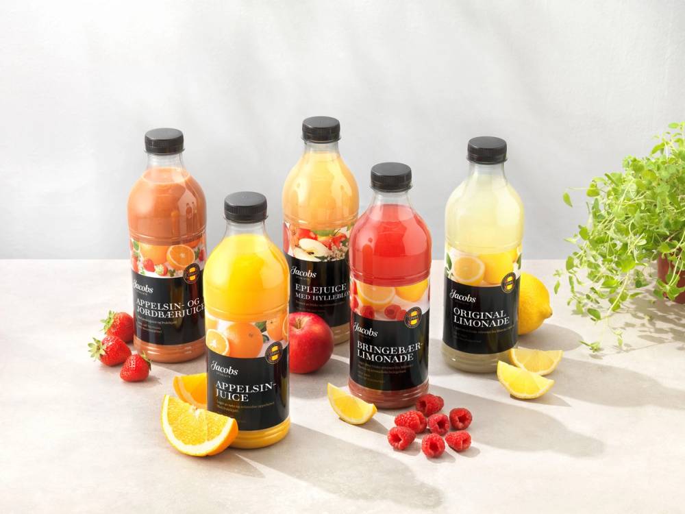 Fem flasker med ulike typer juice, omgitt av frukt som sitron, appelsin, jordbær og rips, på et hvitt bord med en grønn plante til høyre.