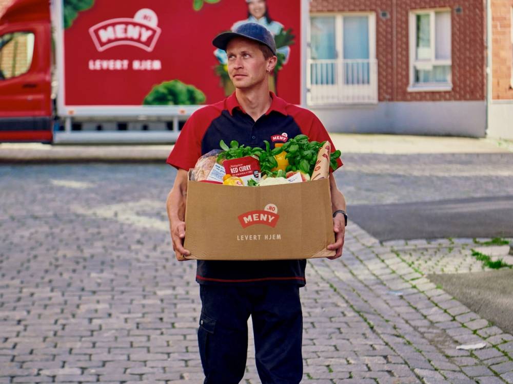 En person i rød og blå uniform holder en stor pose med mat fra Meny, foran en rød leveringsbil med Meny-logoet, på en gate med bygninger i bakgrunnen.