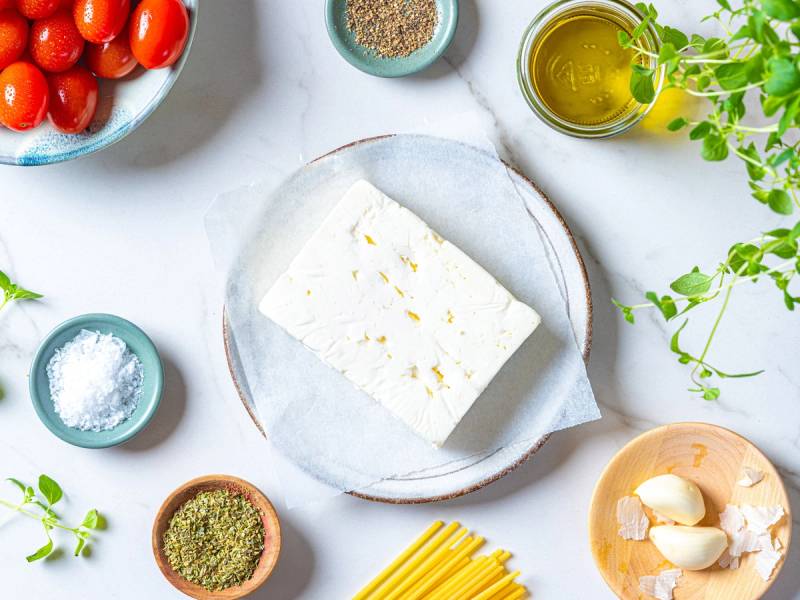 Fetaost på en hvit tallerken omgitt av ulike krydder, olivenolje og tomater på en hvit bakgrunn