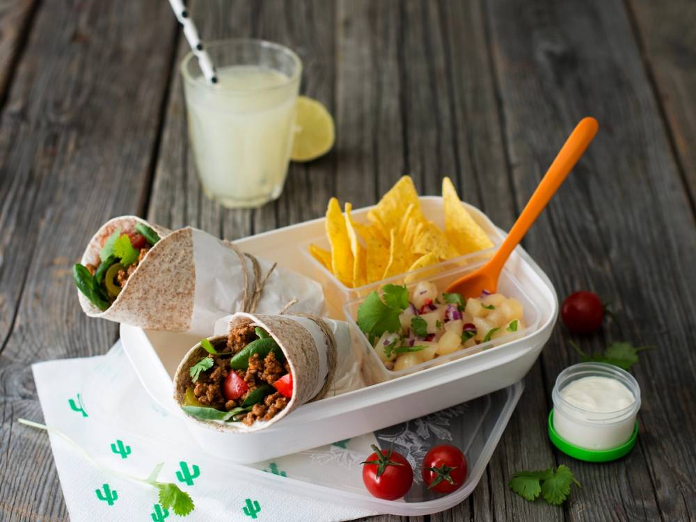 To tacowraps med grønnsaker og kjøtt, servert med en skål med chips, tomater, en sitron, en drink og en liten kopp med saus på et trebord.