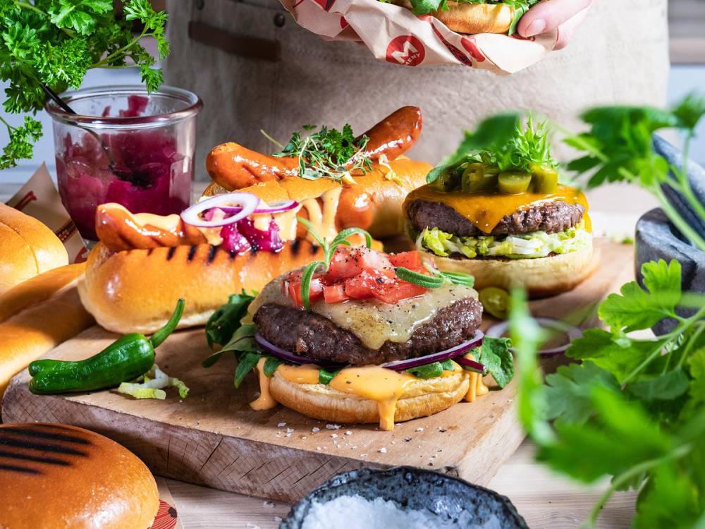 Et utvalg av burgere med pølser, grønnsaker og brød på et serveringsbrett, omgitt av friske urter og tilbehør.