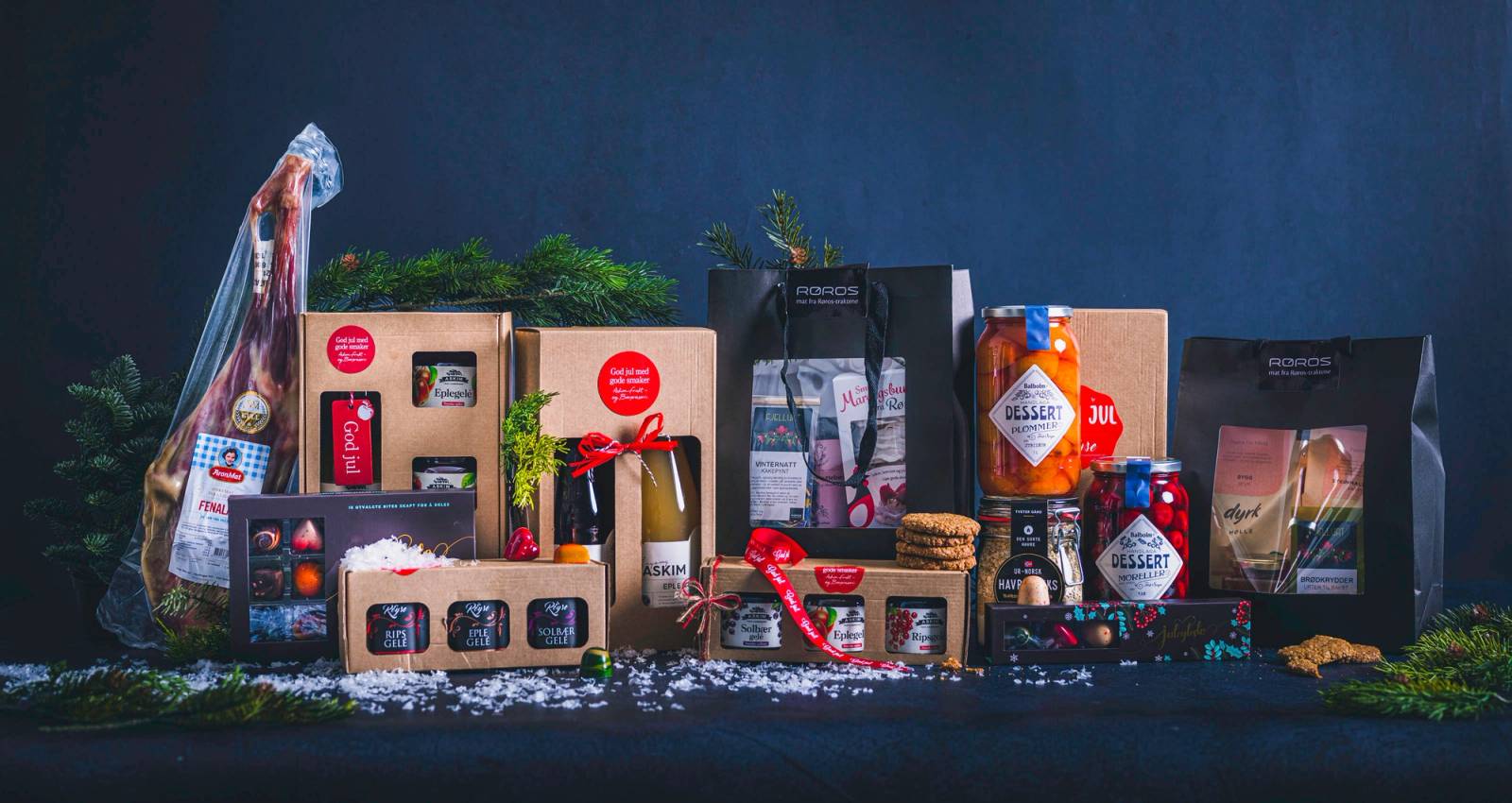 Ulike matgaver og gaveesker er plassert på et bord med en mørk bakgrunn, dekorert med granbar og snø, klar til å gis bort.