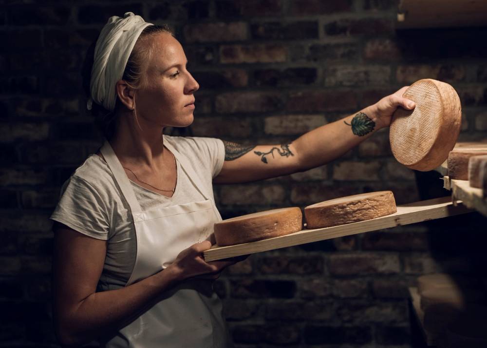 En person med blondt hår og tatoveringer på armen holder en stor skive brød i en bakeri- eller kafémiljø med en murvegg i bakgrunnen.