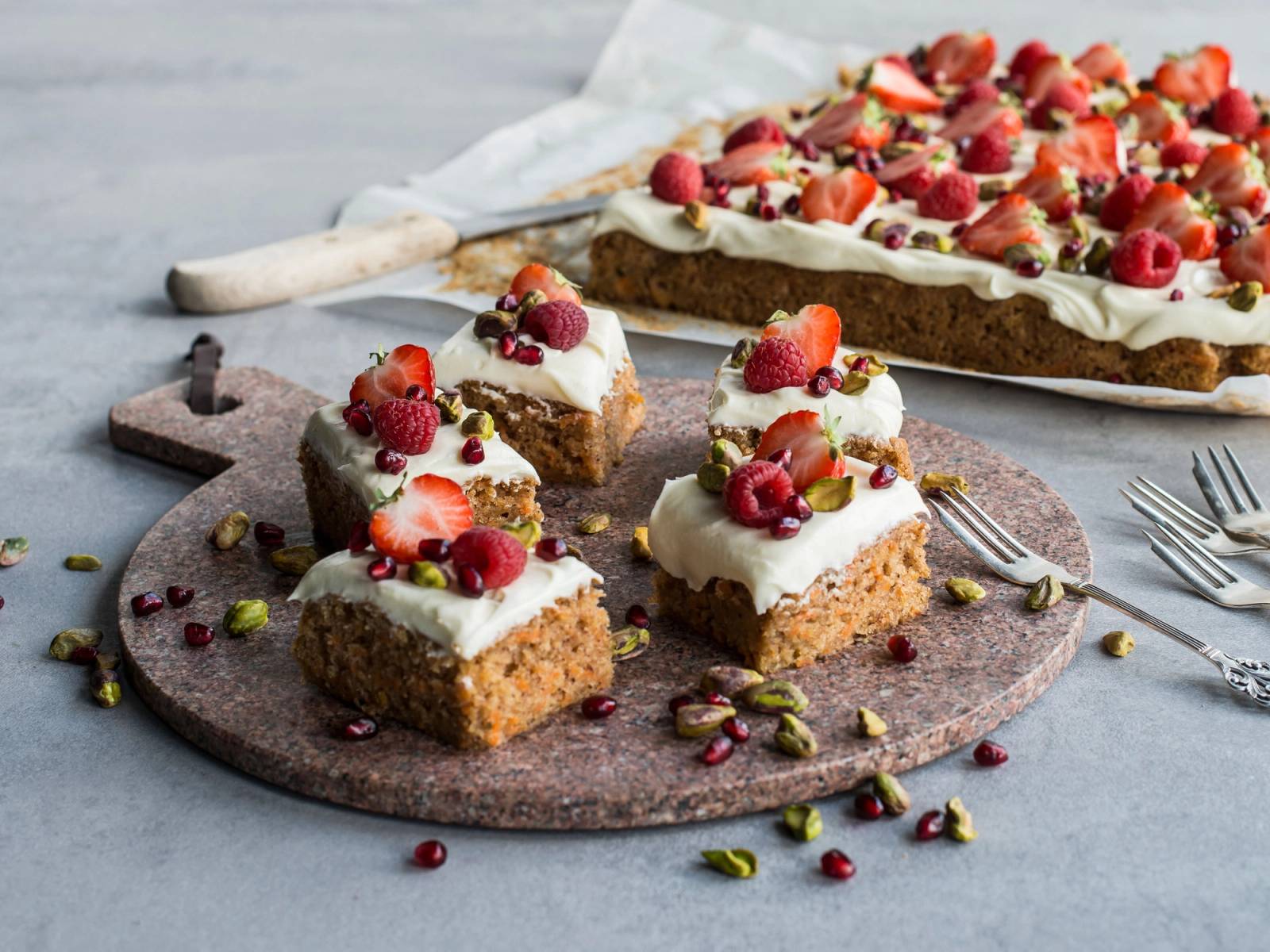 Seks små kaker med hvit krem og bær på en rund steinplate, med en større kake i bakgrunnen pyntet med bær og krem.