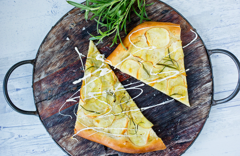 Hvit pizza med ost og rosmarin på en rund treplate med håndtak, garnityr av frisk urter ved siden av.