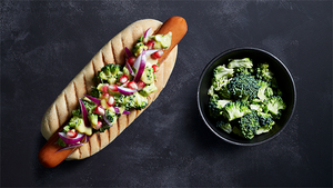 En hotdog med brokkoliblanding og rød løk, ved siden av en skål med fersk brokkoli på en svart bakgrunn.