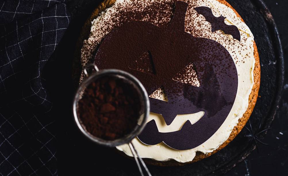 En Halloween-kake med et gresskaransikt som blir dekorert med melis, sett ovenfra.